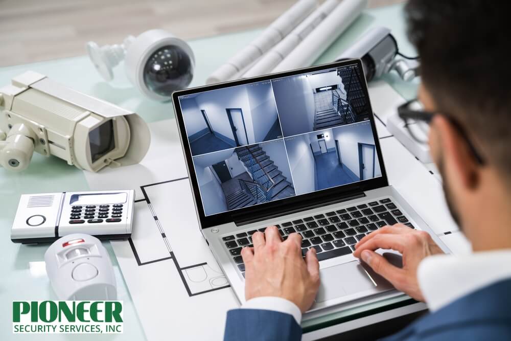 A security technician setting up a small business CCTV system on a laptop, showing multiple interior live feeds, surrounded by cameras and blueprints.